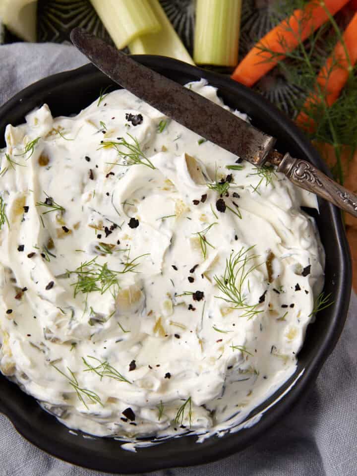 A black bowl filled with creamy dill pickle dip, garnished with fresh dill and cracked pepper; a vintage knife rests on the bowl. Celery, carrot sticks, and crackers are arranged nearby on a gray cloth.