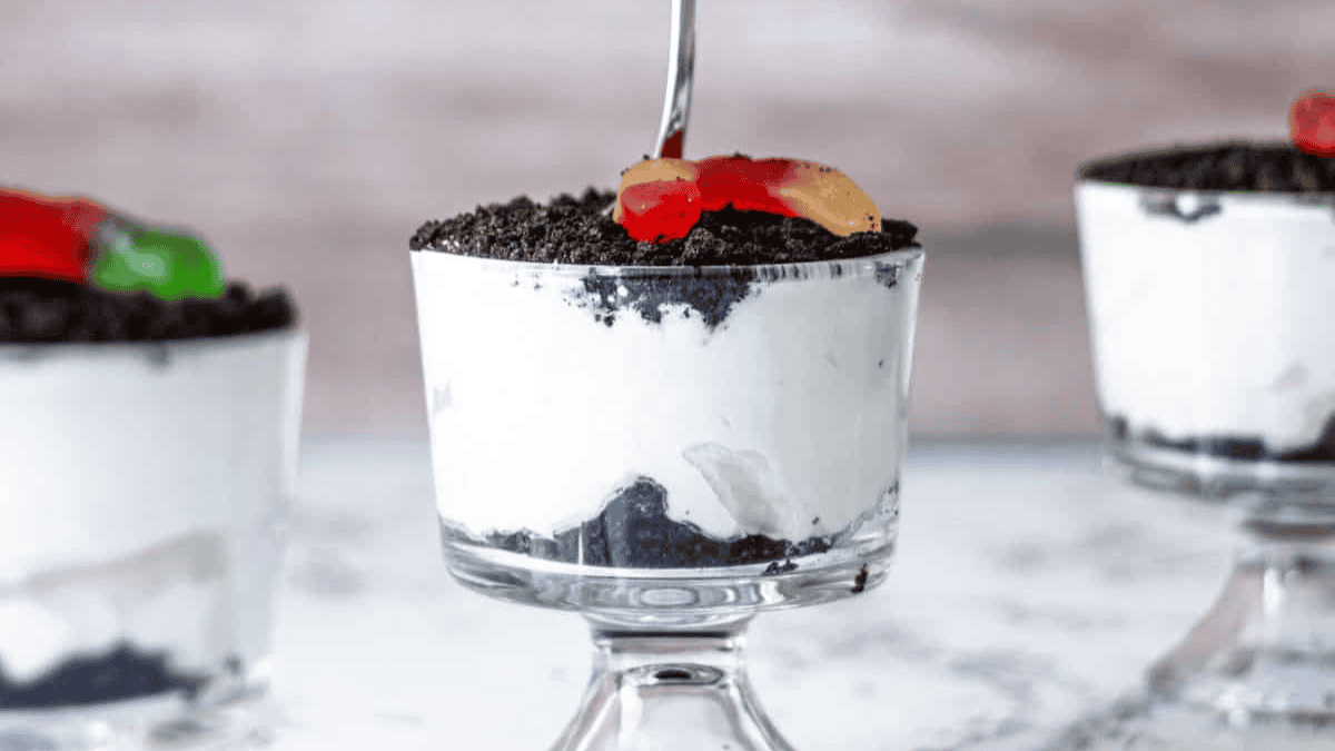 A glass dessert cup filled with layers of crushed chocolate cookies and creamy white pudding, topped with more cookie crumbs and colorful gummy worms. Two similar cups are visible in the background.