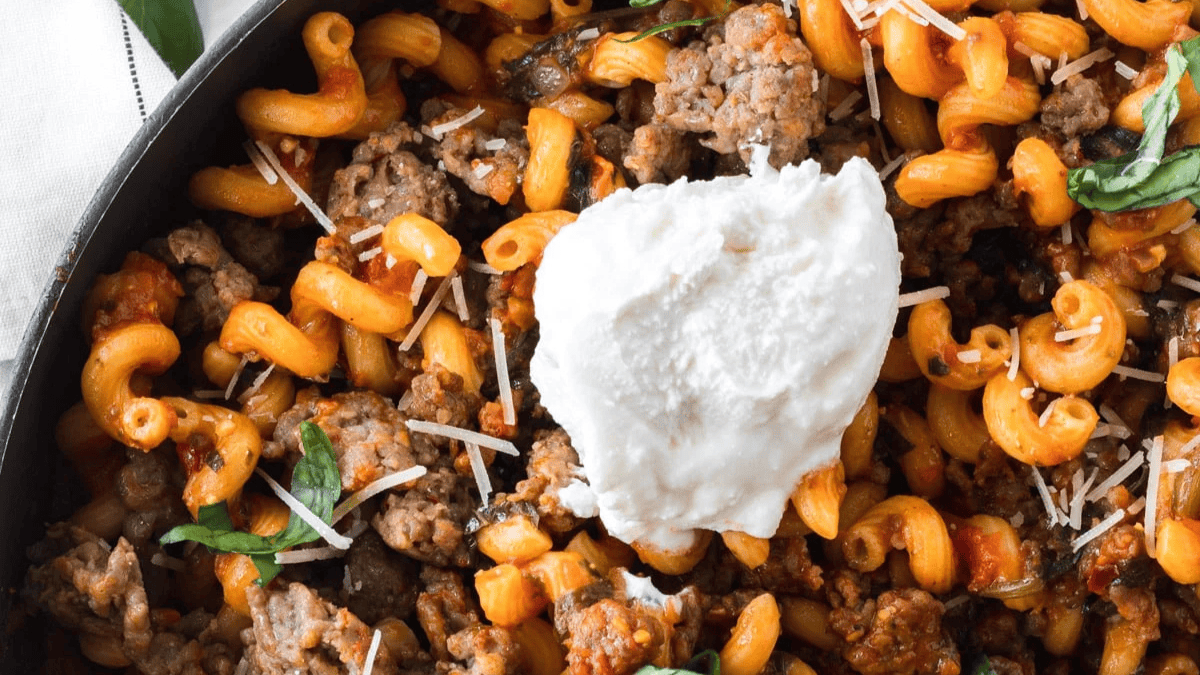A close-up of cooked cavatappi pasta with ground sausage, tomato sauce, fresh basil, grated cheese, and a dollop of ricotta in the center.