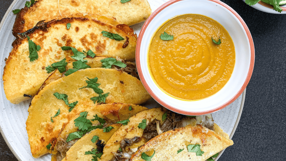 A plate of crispy tacos filled with shredded meat, garnished with fresh cilantro, is served with a bowl of smooth orange dipping sauce.