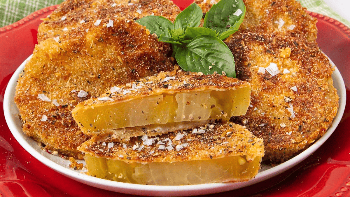 Golden brown fried green tomato slices arranged on a white plate, garnished with fresh basil leaves and sprinkled with flaky sea salt. Two slices are cut open to show the juicy interior.
