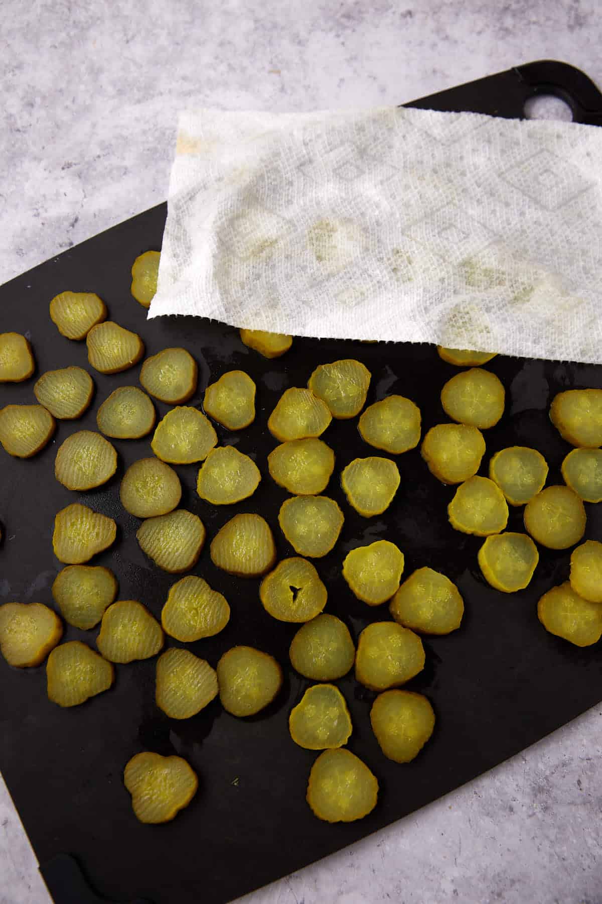 Sliced pickles, perfect for making Southern Fried Pickles, are spread out on a black cutting board, with a paper towel covering part to absorb moisture. The background features a light-colored, textured surface.