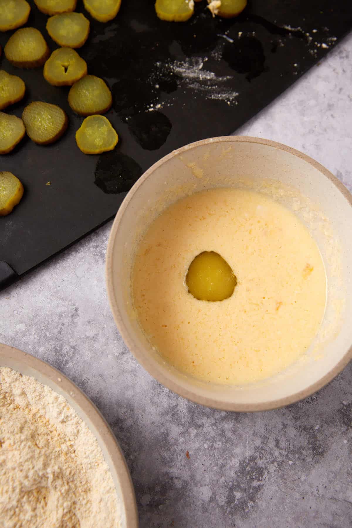 A sliced pickle is dipped in a creamy batter in a bowl for Southern Fried Pickles, with more pickle slices on a dark tray and a plate of flour mixture nearby on a gray countertop.