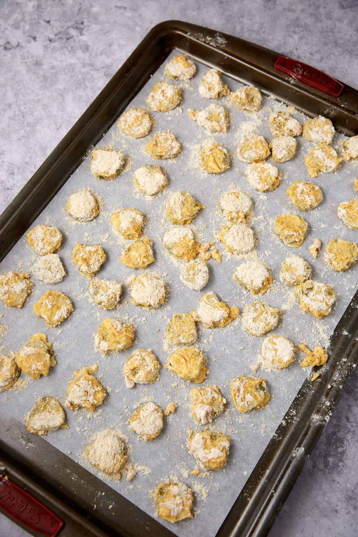 A baking sheet lined with parchment paper holds rows of uncooked, breaded Southern Fried Pickles on a gray countertop, ready to be baked.