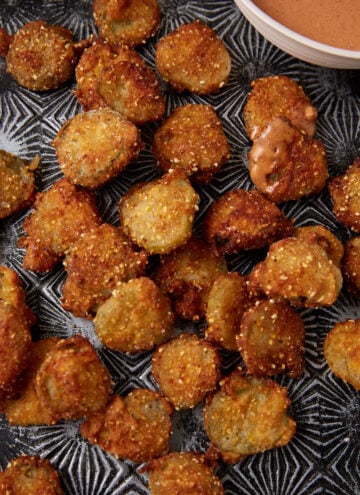 A tray of crispy, golden-brown Southern Fried Pickles sits beside a bowl of creamy dipping sauce. Some pickles have a bit of sauce on them, and crumbs are scattered around on the dark patterned tray.