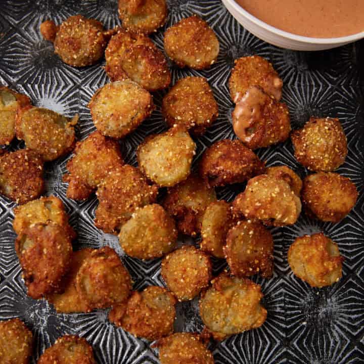 A tray of crispy, golden-brown Southern Fried Pickles sits beside a bowl of creamy dipping sauce. Some pickles have a bit of sauce on them, and crumbs are scattered around on the dark patterned tray.