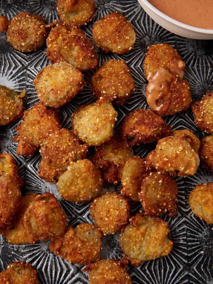 A tray of crispy, golden-brown Southern Fried Pickles sits beside a bowl of creamy dipping sauce. Some pickles have a bit of sauce on them, and crumbs are scattered around on the dark patterned tray.