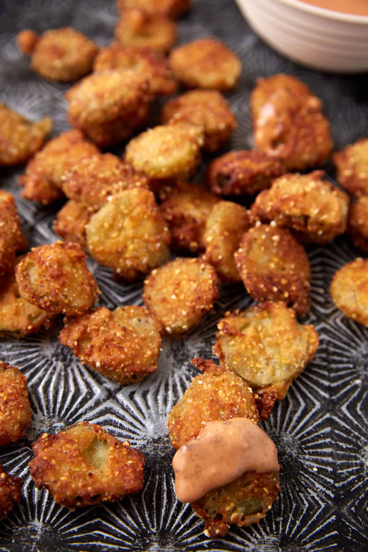 A close-up of crispy Southern Fried Pickles on a textured black and white surface, with one pickle chip dipped in a creamy dipping sauce.