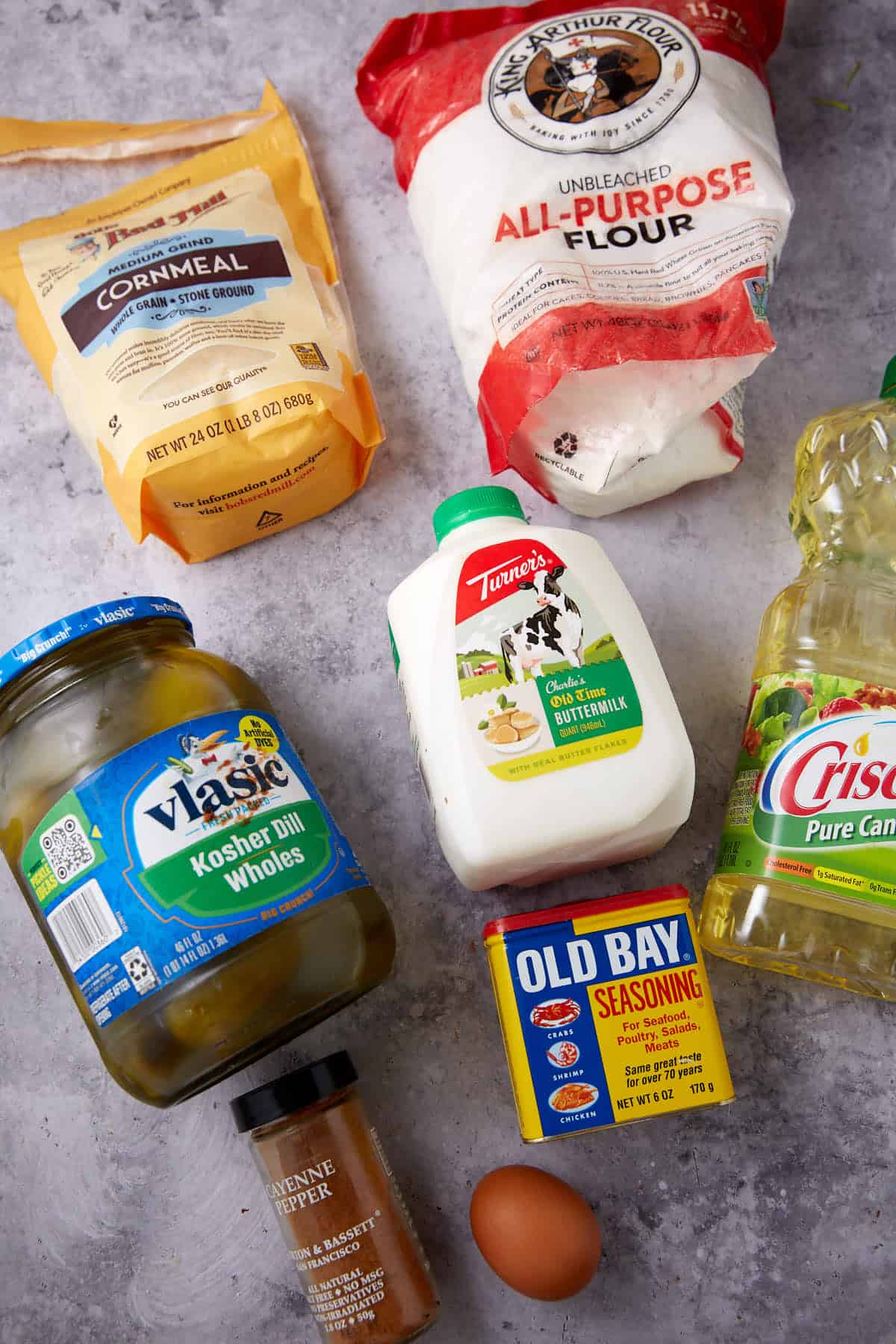 Various cooking ingredients on a grey surface, including cornmeal, all-purpose flour, buttermilk, dill pickles, canola oil, cayenne pepper, Old Bay seasoning, and a single brown egg—everything you need for crispy Southern Fried Pickles.