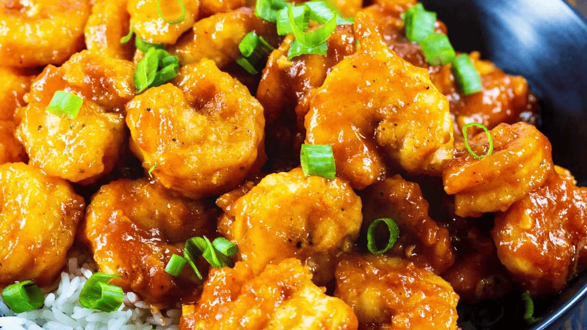 A close-up of saucy, golden-brown shrimp garnished with chopped green onions, served over a bed of white rice in a black bowl.