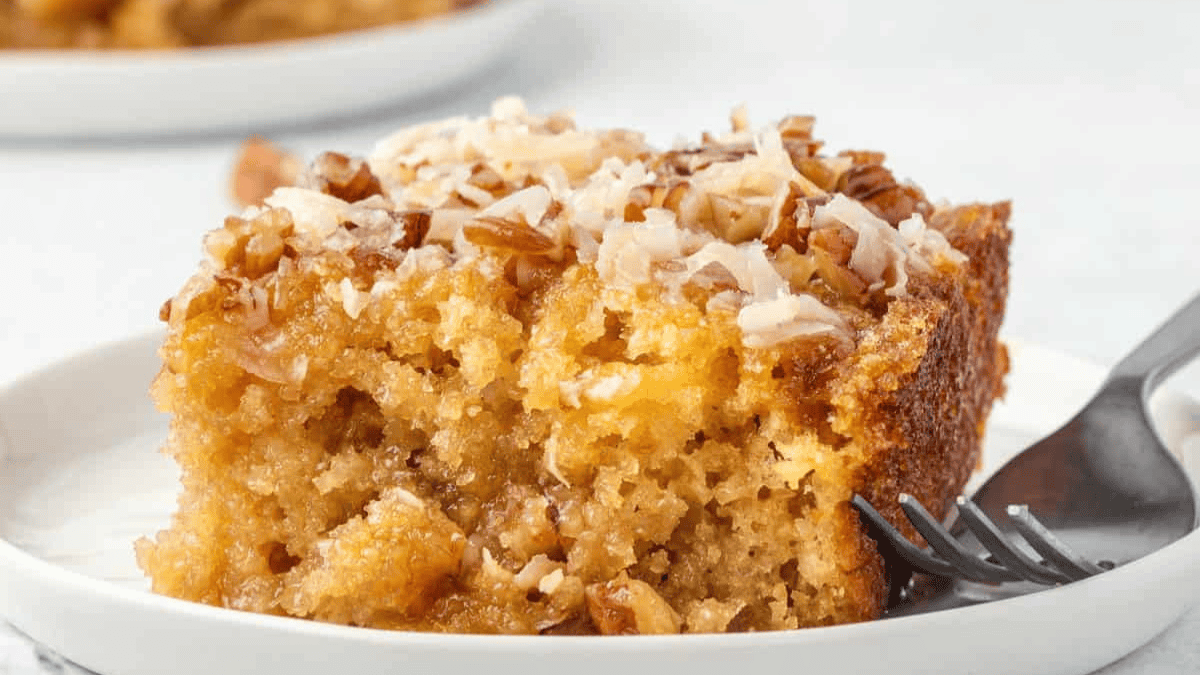 A close-up of a moist slice of cake topped with chopped nuts and shredded coconut on a white plate, with a fork beside it. Another plate with more cake is visible blurred in the background.