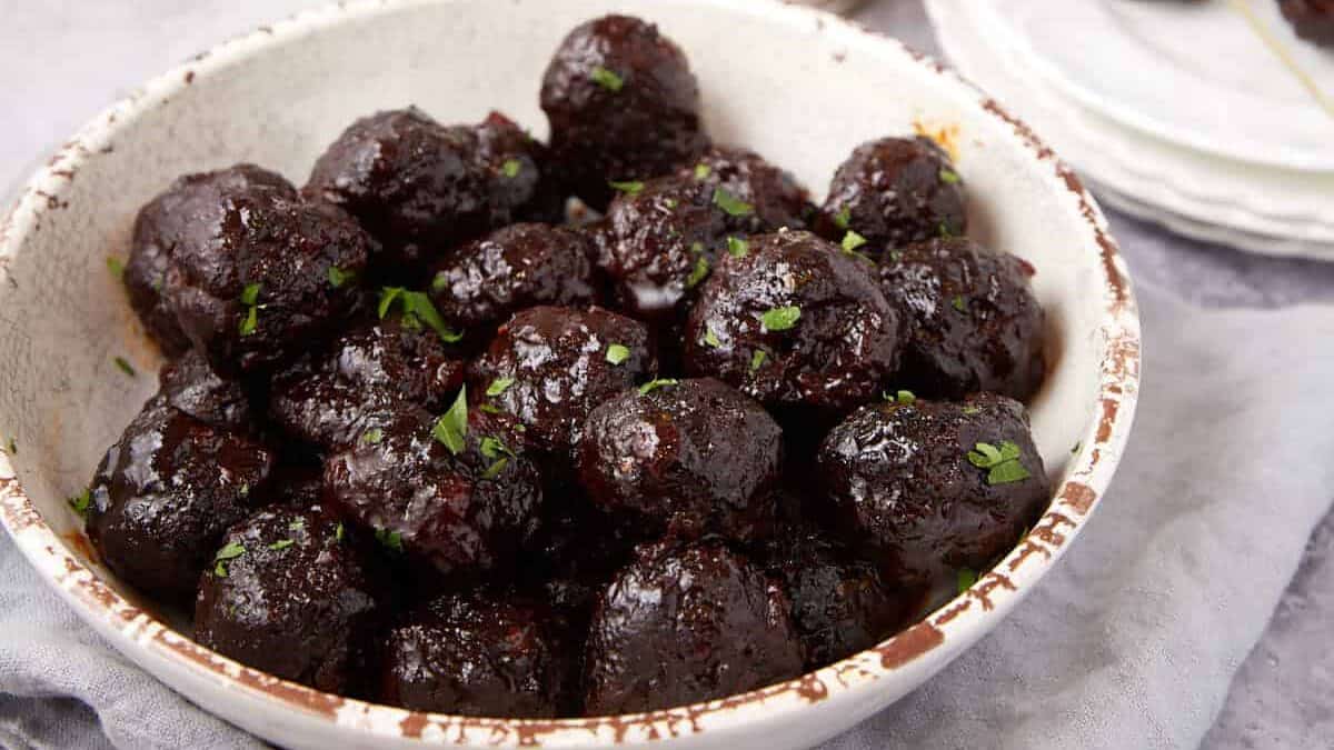 A hand holds a toothpick with a Grape Jelly Meatball above a bowl filled with more glazed meatballs, garnished with herbs. Plates and extra toothpicks are in the background on a gray surface.