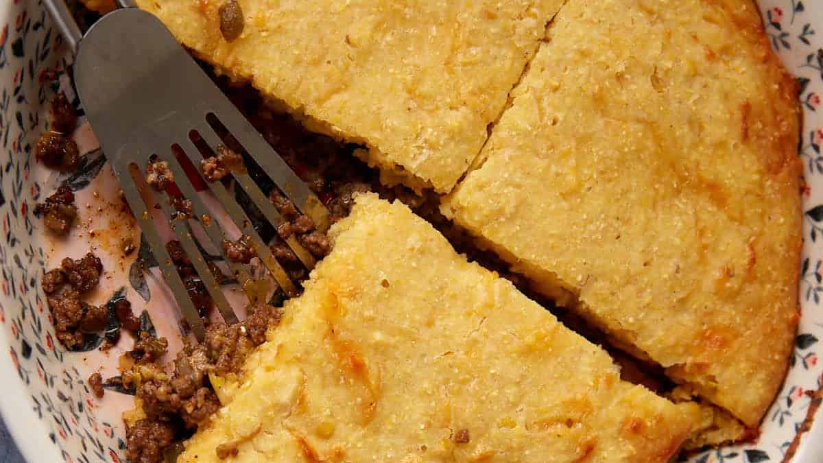 A round dish filled with a baked savory hamburger pie featuring a golden-brown crust and a visible meat filling. A triangular slice has been cut and a spatula is lifting it out. The dish has a floral pattern on its rim, and part of the pie is served on a plate nearby.
