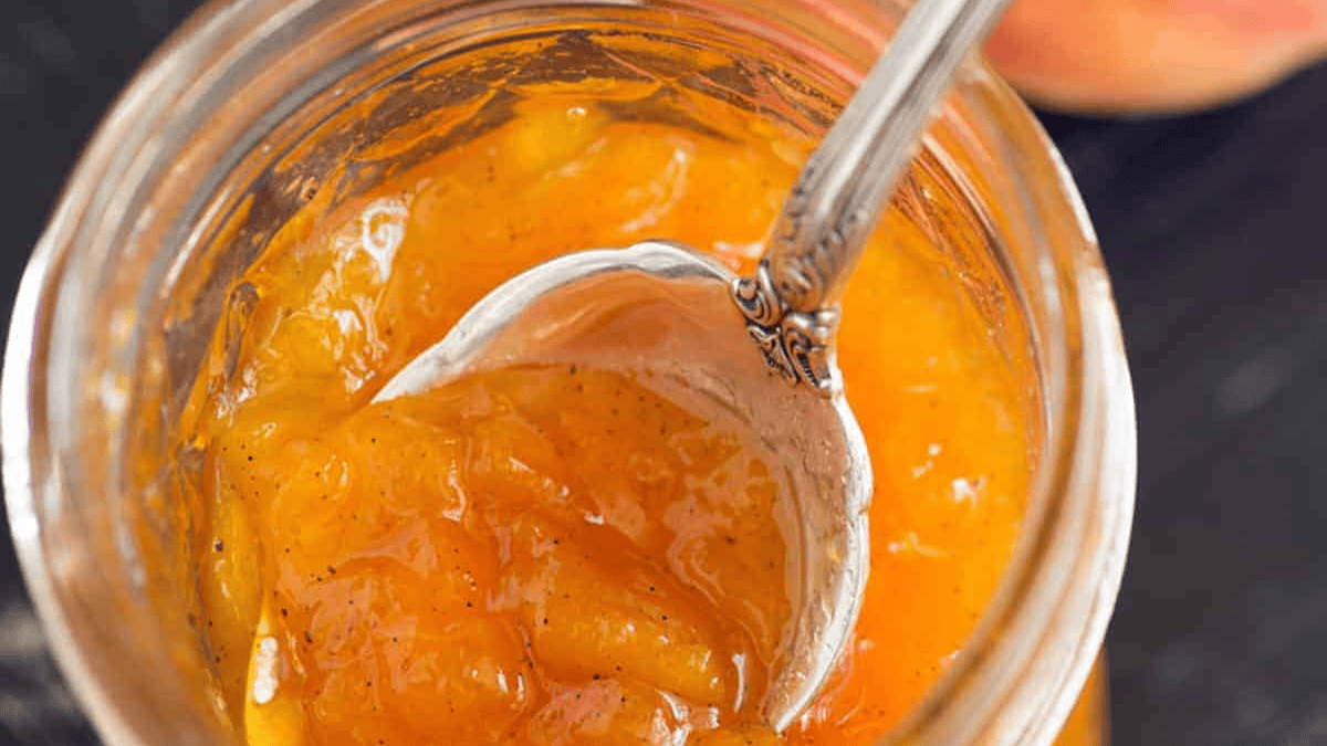 A close-up view of a jar filled with orange-colored peach jam, with a spoon inside scooping out some of the chunky preserve.