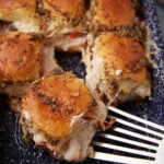 A close-up of golden-brown Hot Brown Sliders filled with turkey and cheese, topped with herbs and poppy seeds, being lifted from a blue speckled baking dish with a metal spatula.