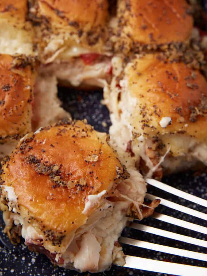 A close-up of golden-brown Hot Brown Sliders filled with turkey and cheese, topped with herbs and poppy seeds, being lifted from a blue speckled baking dish with a metal spatula.