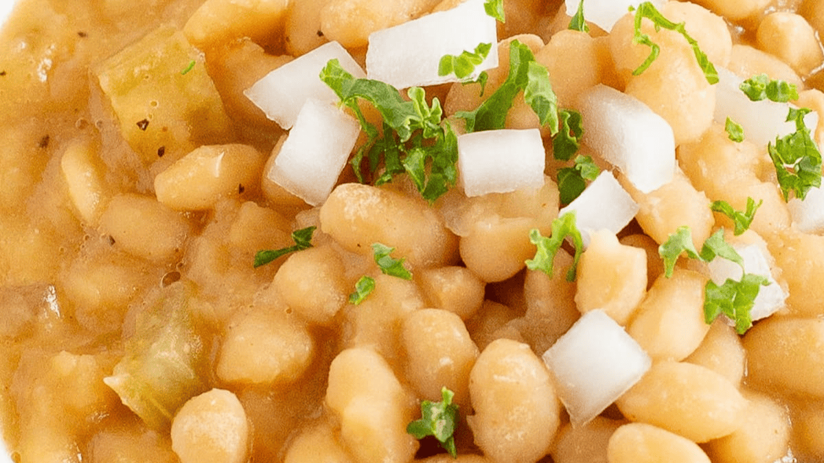 Close-up of baked beans garnished with chopped white onions and fresh parsley. The beans appear to be in a thick, creamy sauce with small pieces of green vegetables mixed in.