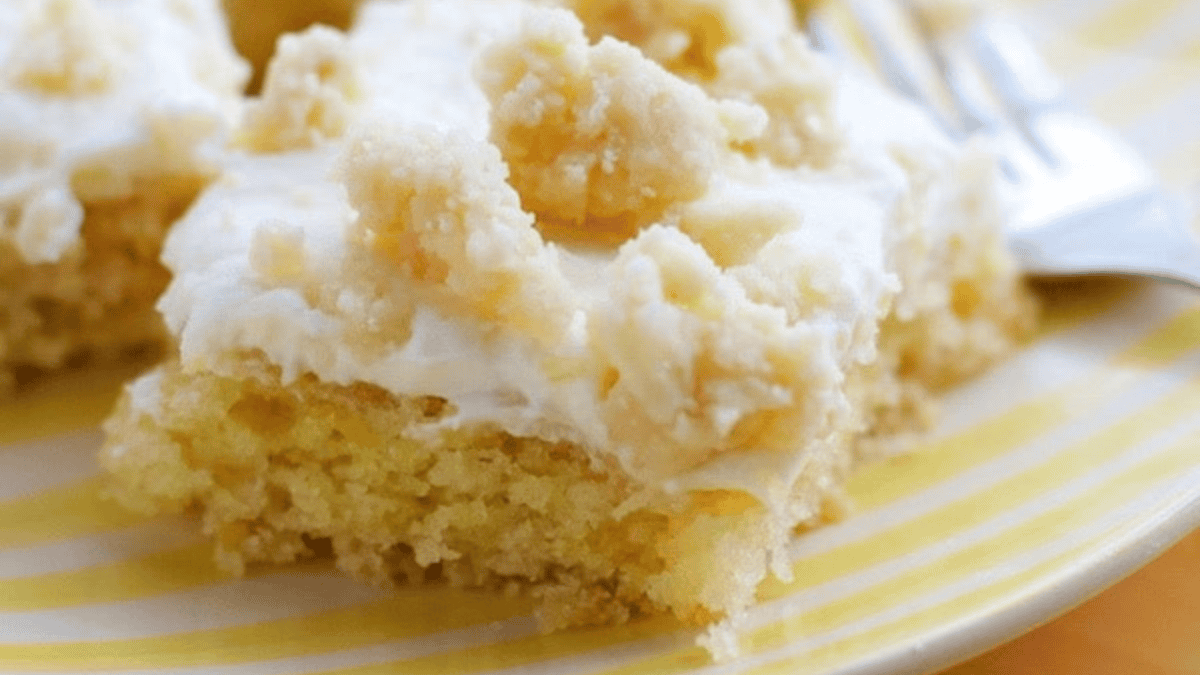 A close-up of a crumbly dessert bar with a yellow cake base, thick layer of creamy white frosting, and crumb topping, served on a yellow striped plate with a fork in the background.