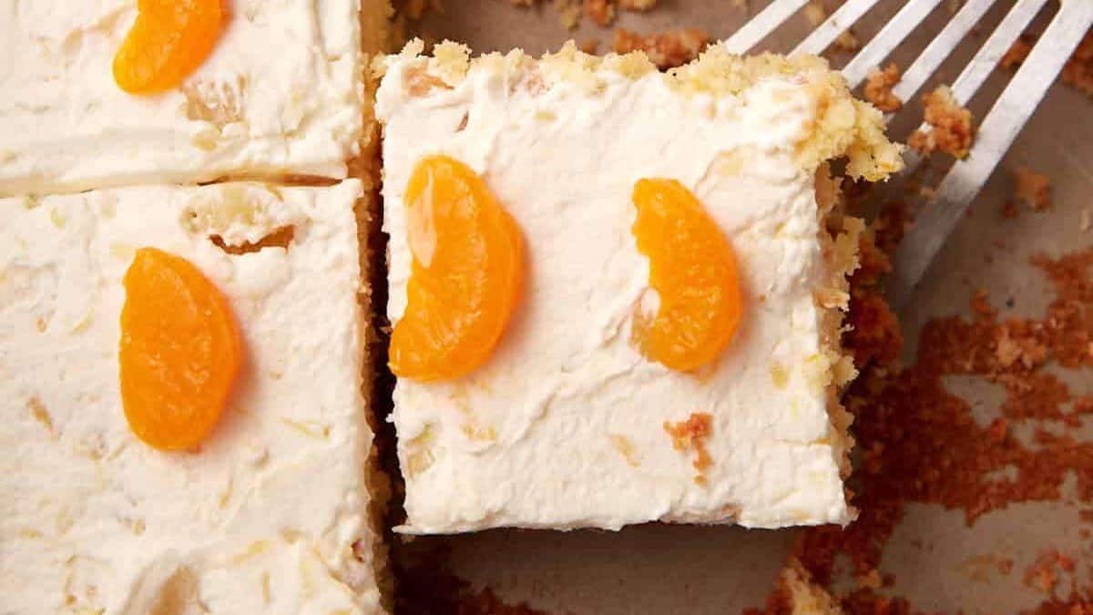 A square piece of cake with creamy white frosting and two orange mandarin slices on top is being lifted from a tray with a metal spatula—one of these cakes might be yours. Crumbs and more cake pieces are visible in the background.