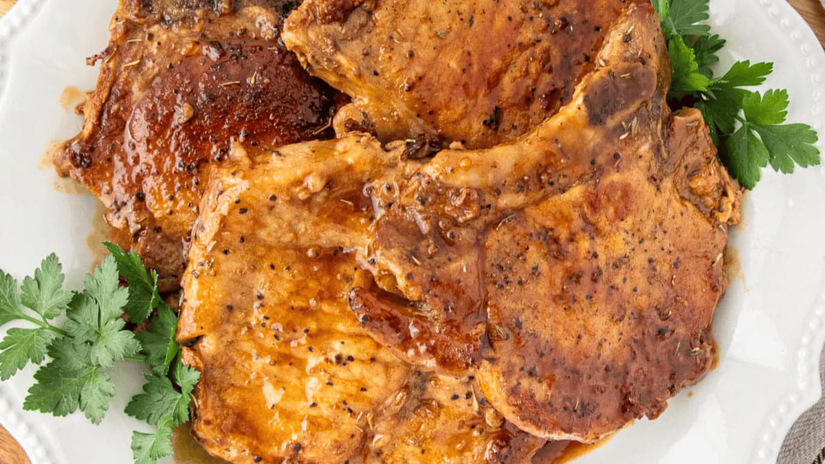 Three cooked pork chops with a golden-brown glaze are served on a white plate, garnished with fresh parsley on the side.