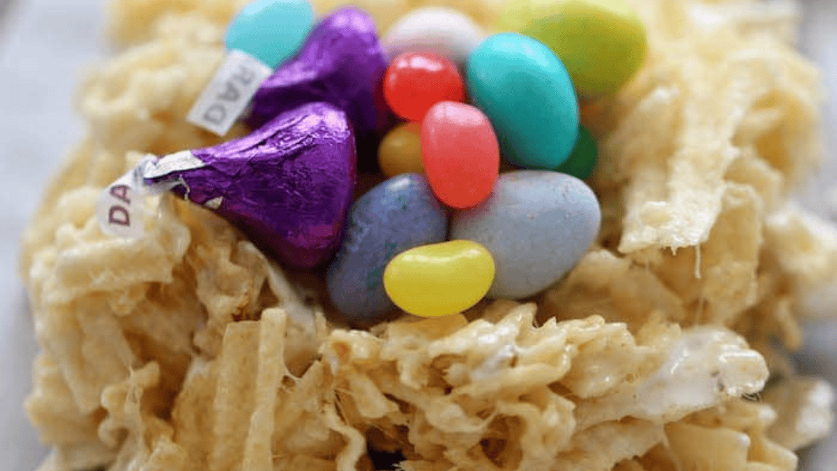 A close-up of a treat made from crunchy, light-colored cereal, topped with colorful jellybeans, candy-coated chocolate eggs, and two purple-wrapped chocolate kisses—you might judge these American comfort foods, but they’re delightfully fun.