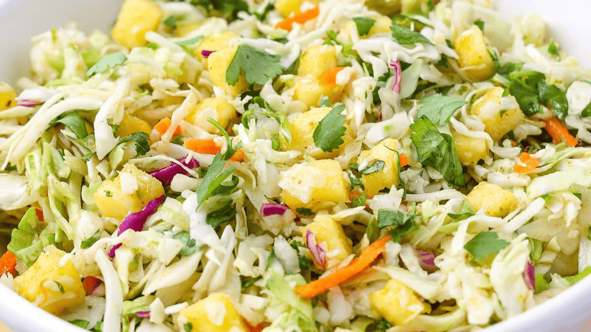 A close-up of a colorful coleslaw salad with shredded cabbage, carrots, red cabbage, fresh cilantro, and small chunks of pineapple in a white bowl.