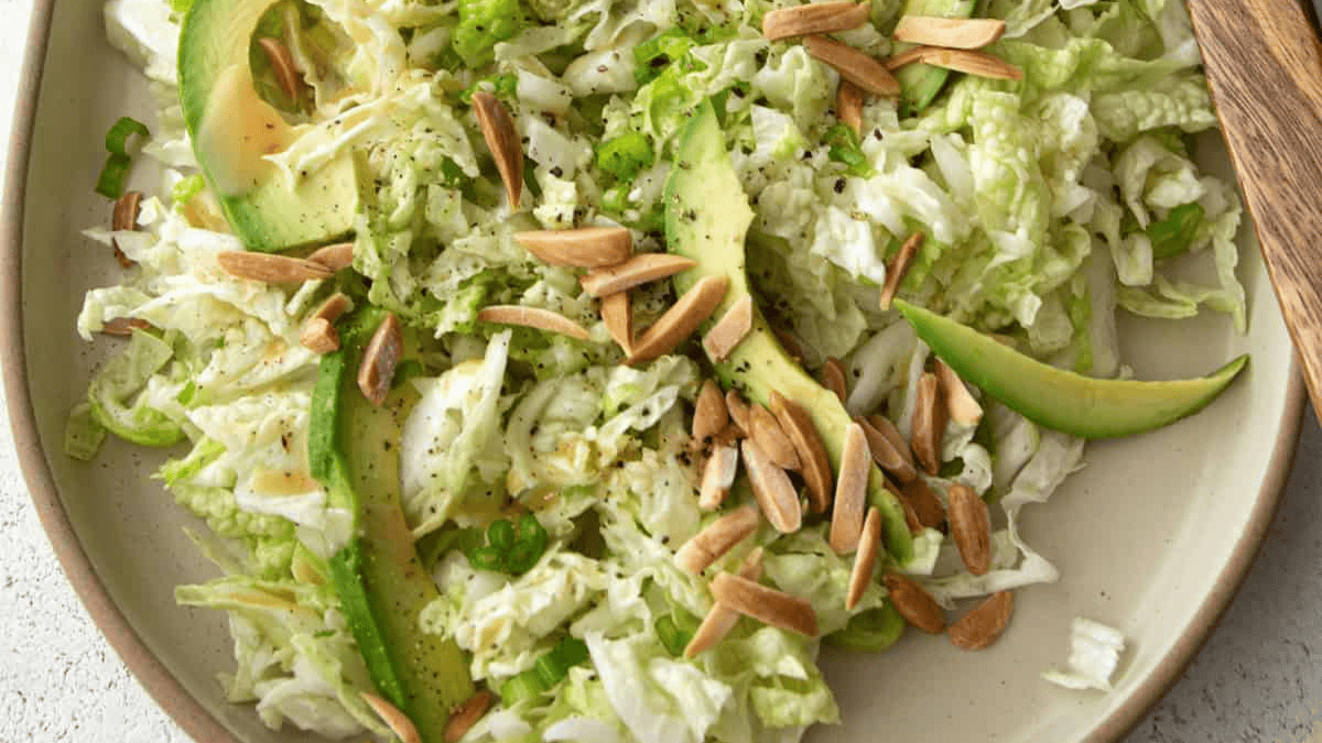 A plate of shredded cabbage salad topped with avocado slices, toasted almond slivers, chopped green onions, and freshly ground black pepper.