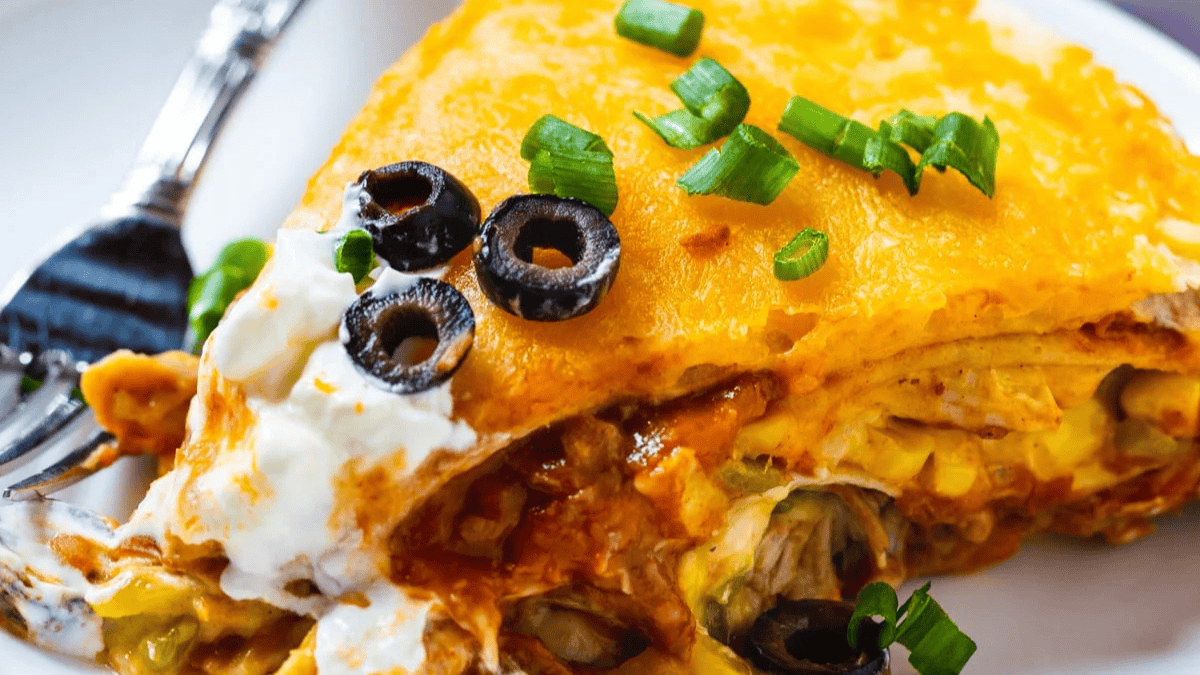 A close-up of a cheesy enchilada topped with sliced black olives, chopped green onions, and sour cream on a white plate with a fork.