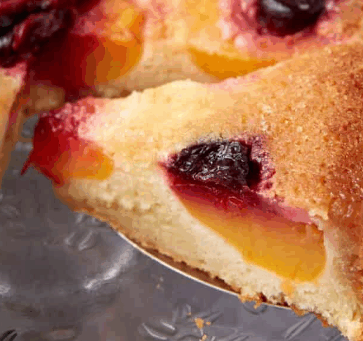 A close-up of a fruit-studded cake, showing a slice being lifted. The cake is golden-brown with pieces of yellow peaches and dark red plums baked into the top.