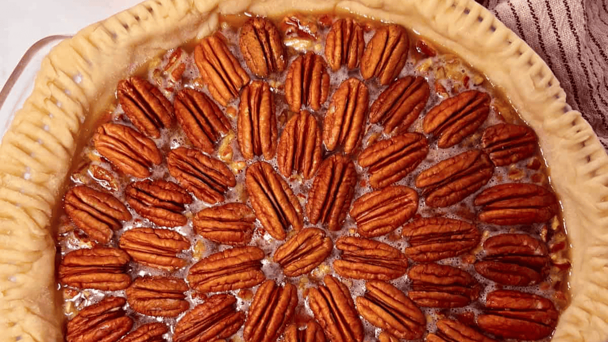 A close-up of a pecan pie with whole pecans arranged in a circular pattern on top of a golden, crimped pie crust. The filling is glossy and caramelized.