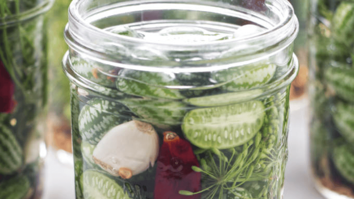 A glass jar filled with pickled cucamelons, garlic cloves, red chili, and fresh dill, ready for preservation.