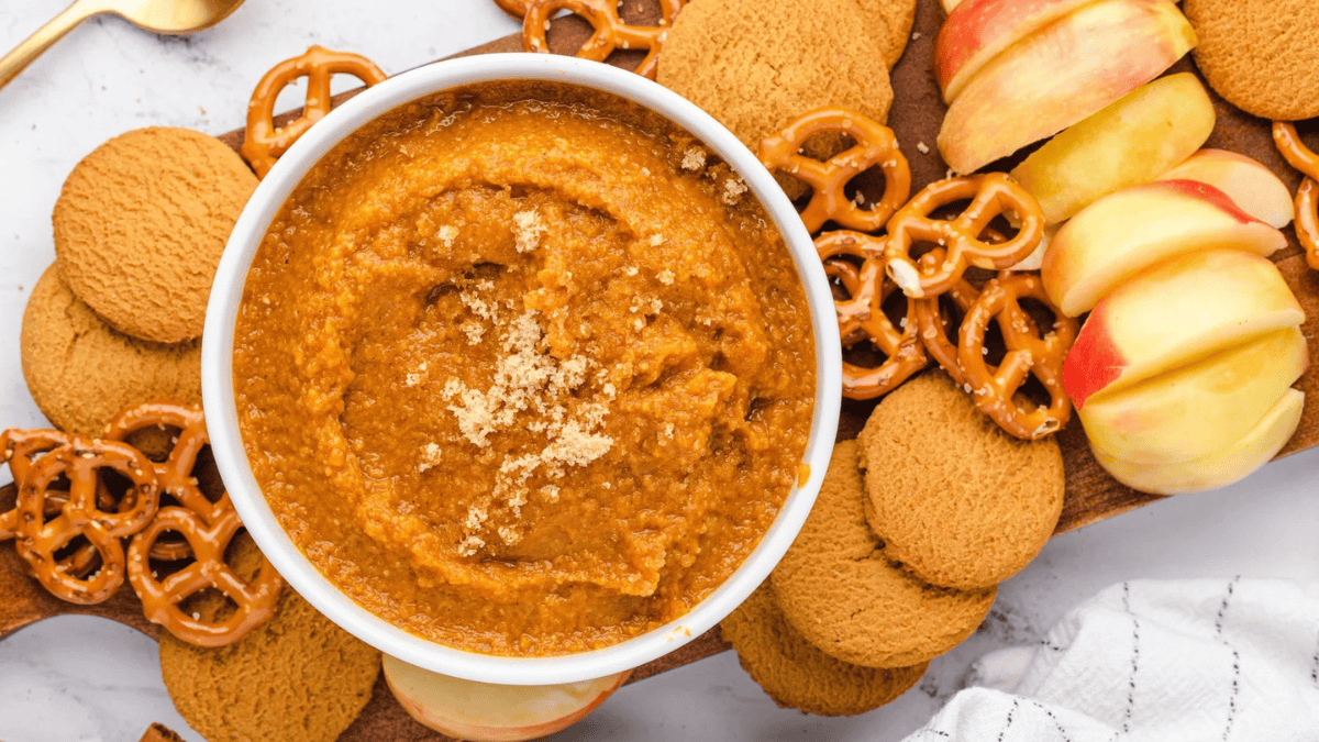 A bowl of brown pumpkin dip is surrounded by ginger cookies, pretzels, and sliced apples on a wooden board. Some crumbs are sprinkled on top of the dip—just one of those American comfort foods you might judge before taking a bite.