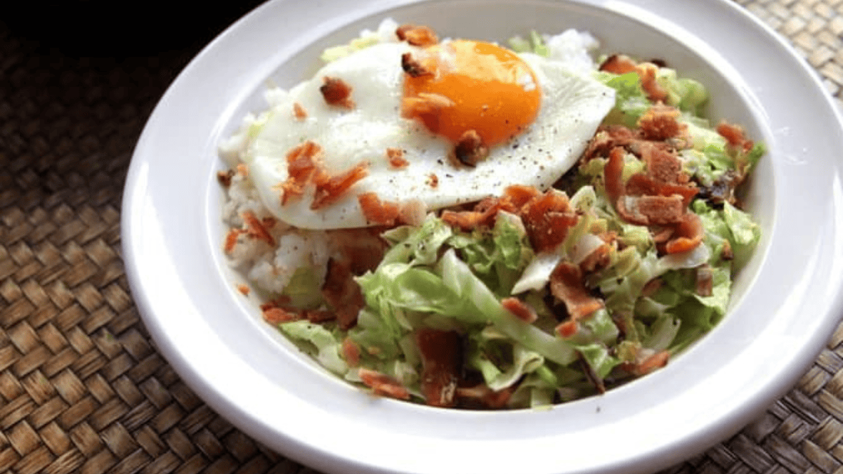 A white bowl filled with rice, sautéed cabbage, crumbled bacon, and topped with a sunny-side-up egg, placed on a woven placemat.