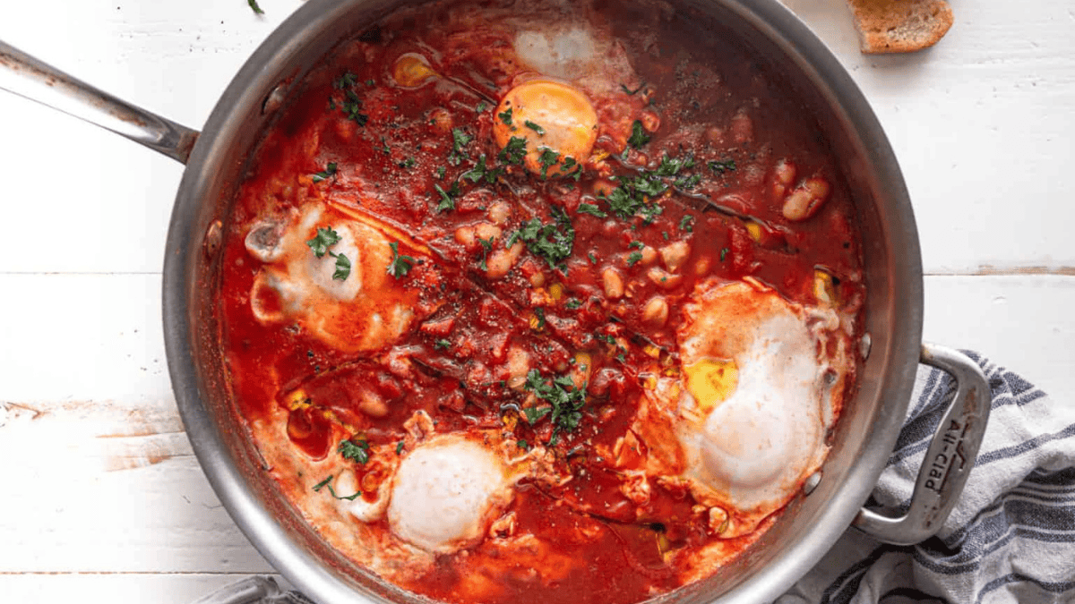 A pan filled with a tomato-based stew featuring poached eggs, white beans, and fresh herbs, with a slice of bread and a striped napkin nearby on a white surface.