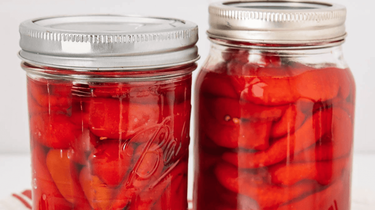 Two glass jars filled with red pickled peppers in brine, sealed with metal lids, are placed side by side on a white surface.