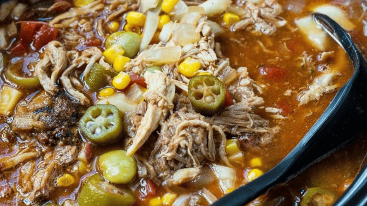 A close-up of Brunswick stew featuring shredded meat, sliced okra, corn, tomatoes, onions, and a rich, reddish broth, with a black serving spoon partially visible.