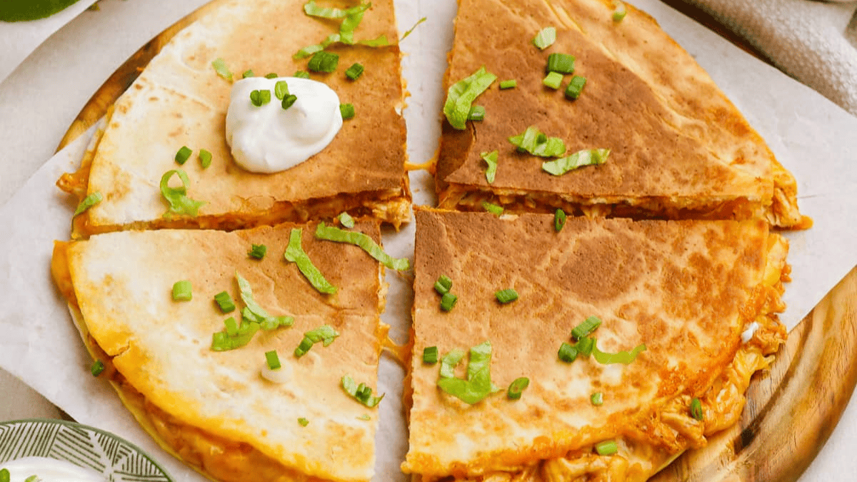 A cheese quesadilla cut into six slices, topped with chopped green onions, shredded lettuce, and a dollop of sour cream on one slice, served on parchment paper.