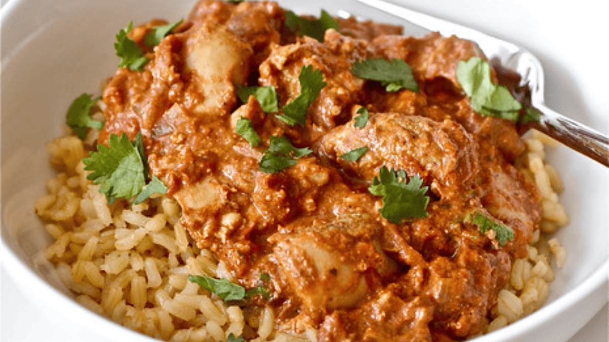A bowl of brown rice topped with creamy, reddish-orange curry chicken, garnished with fresh cilantro leaves, with a spoon on the side.