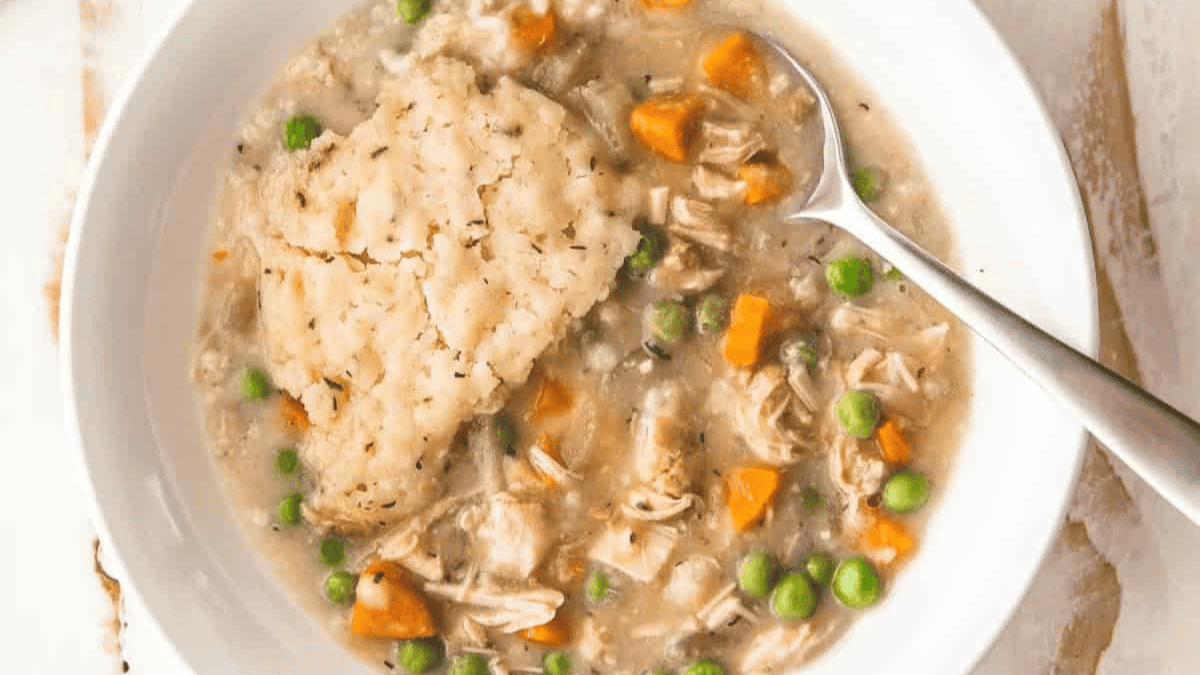 A bowl of creamy chicken and vegetable stew with peas and carrots, topped with a large, fluffy herb dumpling. A spoon rests inside the bowl.