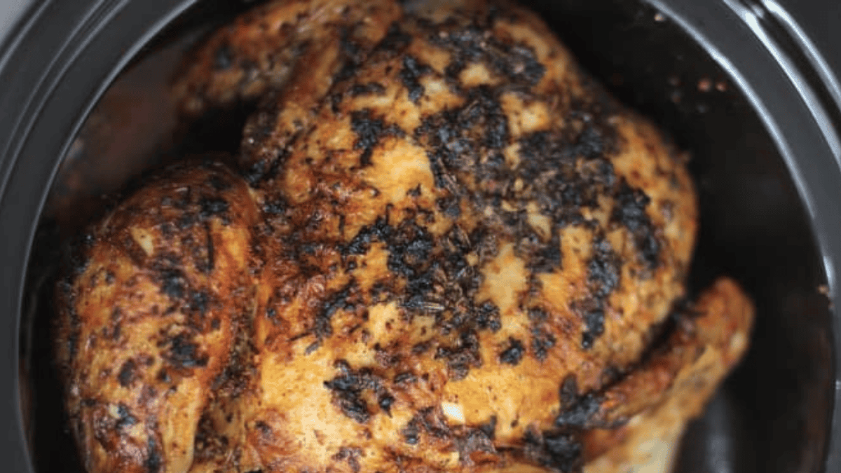 A cooked whole chicken with crispy, browned skin and visible seasoning, resting in a black slow cooker or pot.