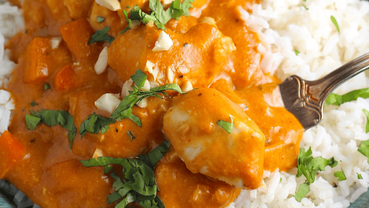 Close-up of creamy orange chicken curry with chopped herbs and nuts, served over a bed of white rice. A fork rests in the curry, and fresh cilantro is sprinkled on top.