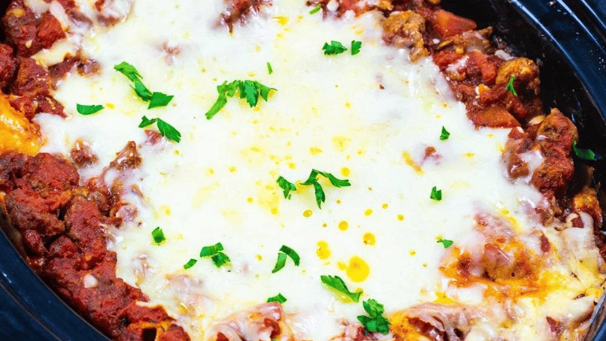A close-up of cheesy baked lasagna in a black slow cooker, topped with melted cheese, tomato sauce, ground meat, and garnished with chopped fresh parsley.