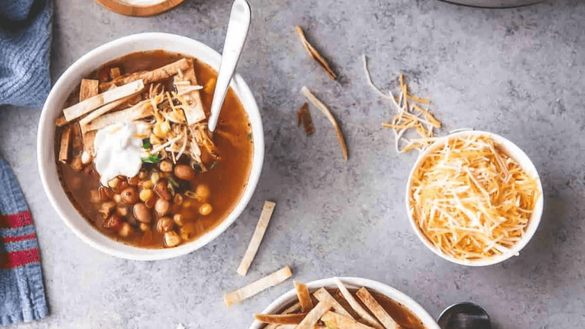 A bowl of tortilla soup topped with shredded cheese, tortilla strips, and sour cream sits on a gray surface beside a small bowl of shredded cheese and scattered tortilla strips.
