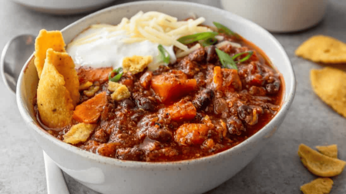 A bowl of chili topped with shredded cheese, sour cream, chopped green onions, and corn chips, with more corn chips scattered nearby on a gray surface.