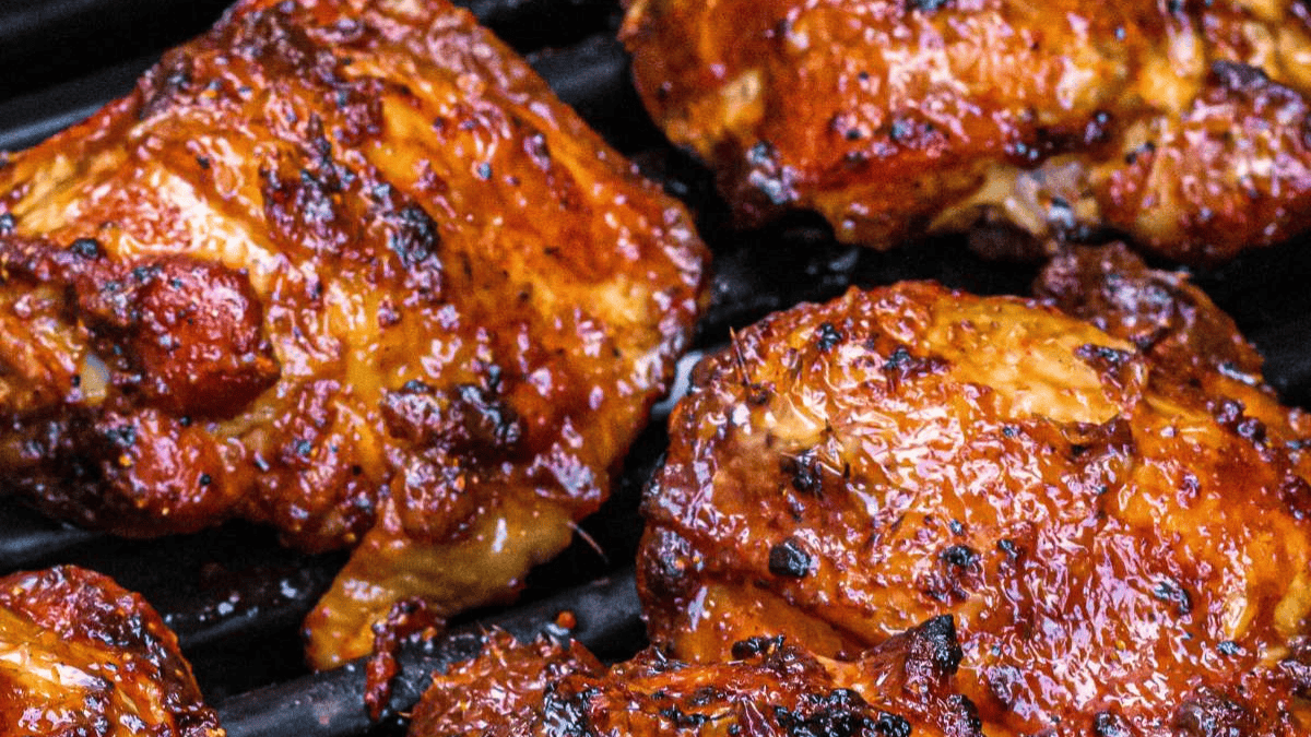 Close-up of several pieces of grilled barbecue chicken with a caramelized, sticky glaze and char marks, resting on a black grill grate.