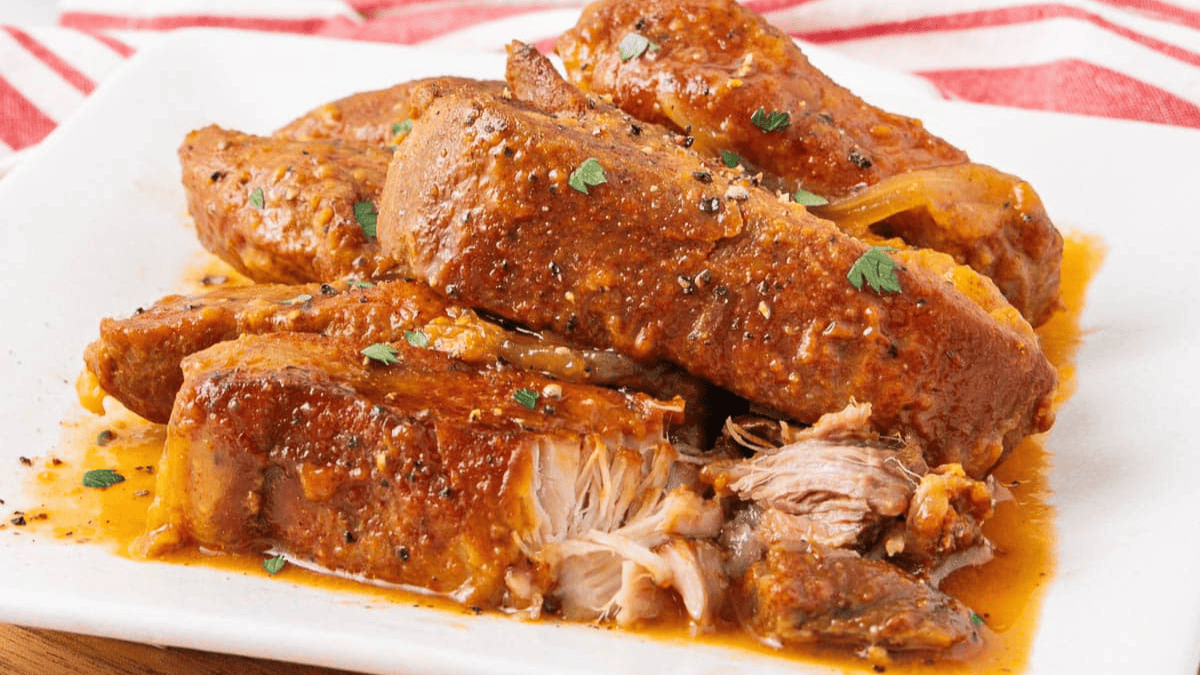 Tender pork roast slices covered in a rich, reddish-brown sauce are served on a white plate, garnished with small pieces of parsley, with a red and white striped cloth in the background.