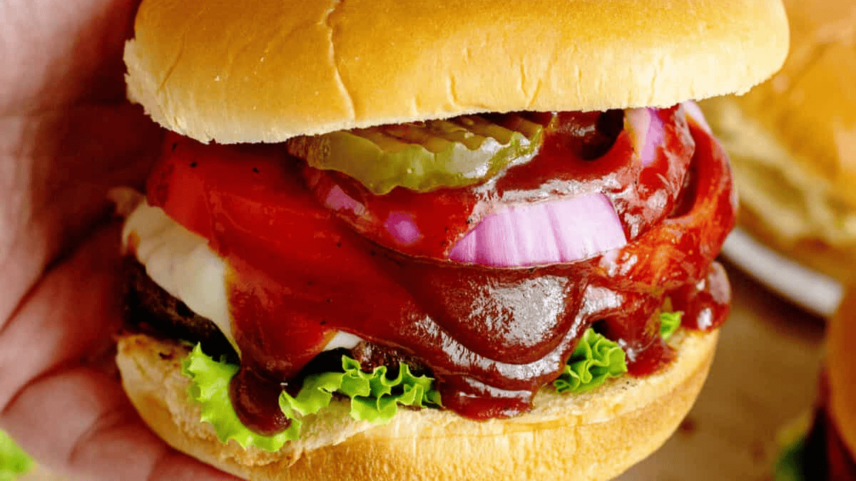 A close-up of a hand holding a hamburger with lettuce, a beef patty, melted cheese, tomato, red onion, pickles, and barbecue sauce in a soft bun.