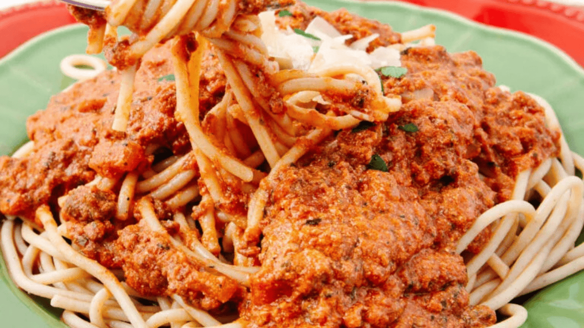 A close-up of spaghetti topped with rich, chunky tomato and meat sauce, garnished with shredded cheese and herbs on a green plate. A fork twirls some pasta—one bite and you might judge these American comfort foods differently!.