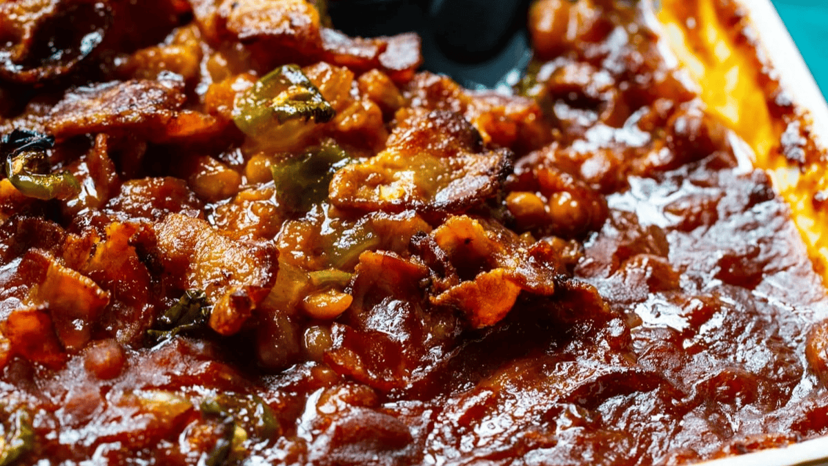Close-up of baked beans topped with crispy bacon pieces and green peppers in a rich, glossy sauce.