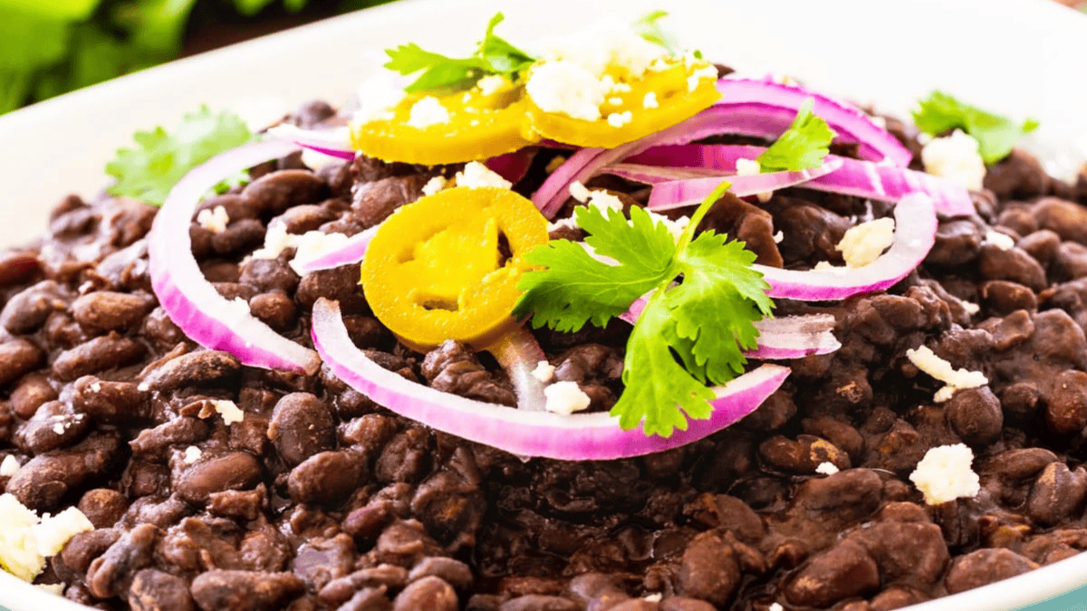 A plate of cooked black beans topped with sliced red onions, pickled jalapeños, fresh cilantro, and crumbled cheese.